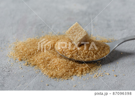 Brown cane sugar crystals and cube of refined sugar in a spoon on grey table. Brown cane sugar crystals and cube of refined sugar in a spoon on grey table. 87096398