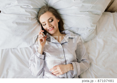 Attractive female with cup of coffee, smartphone and cakes in the bed 87107481