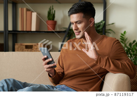 Young man sitting on sofa in the living room and waving his hand to camera of his mobile phone during online conversation Young man sitting on sofa in the living room and waving his hand to camera of his mobile phone during online conversation 87119317