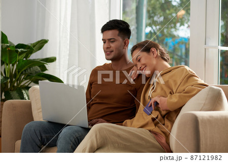 Happy young couple sitting on sofa in the living room and using laptop together, they ordering things online 87121982