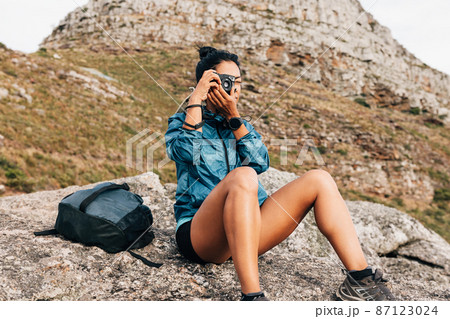 Fit woman in sports clothes with backpack sitting on stone taking photographs on an analog camera Fit woman in sports clothes with backpack sitting on stone taking photographs on an analog camera 87123024