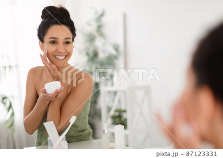 Beautiful millennial woman applying moisturizing face cream, standing in front of mirror after shower at home, copy space Beautiful millennial woman applying moisturizing face cream, standing in front of mirror after shower at home, copy space 87123331