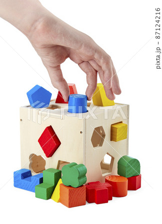 Detail of a wooden children's toy in the hand, developing fine motor skills, consisting of geometric shapes and holes in a cube for them, isolated on a white background with a shadow. Detail of a wooden children's toy in the hand, developing fine motor skills, consisting of geometric shapes and holes in a cube for them, isolated on a white background with a shadow. 87124216