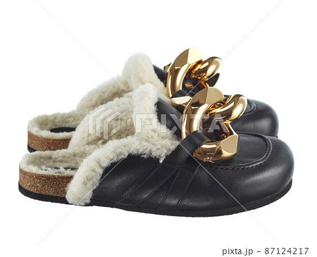 Home slippers with an open heel, made of natural sheepskin, fur inside, black leather outside, decorated with large links of a gold chain isolated on a white background. Side view. Fashionable shoes. 87124217