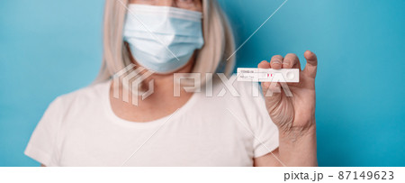 Woman in protective medical mask holding an antigen test with positive result for COVID-19 pandemic posing over bright blue color background Woman in protective medical mask holding an antigen test with positive result for COVID-19 pandemic posing over bright blue color background 87149623