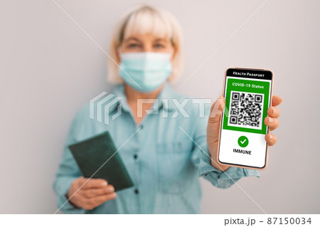 Covid-19 control. 50s caucasian woman in mask holding smartphone with digital green pass and QR code on the screen. Health passport or certificate of immunity. 87150034