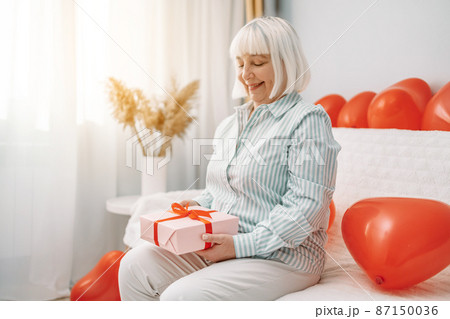 Attractive happy smiling blonde 50s woman holding pink gift box with red ribbon on 14 february sitting on a sofa at home. 87150036