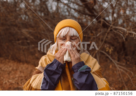 Sick adult woman coughing and blowing her nose in park during autumn season in a cold autumn day. Sick adult woman coughing and blowing her nose in park during autumn season in a cold autumn day. 87150156