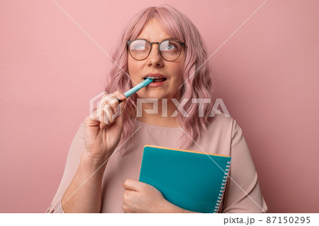 Thoughtful female student wearing glasses holds pen notebook has pensive expression on pink background. Thoughtful female student wearing glasses holds pen notebook has pensive expression on pink background. 87150295