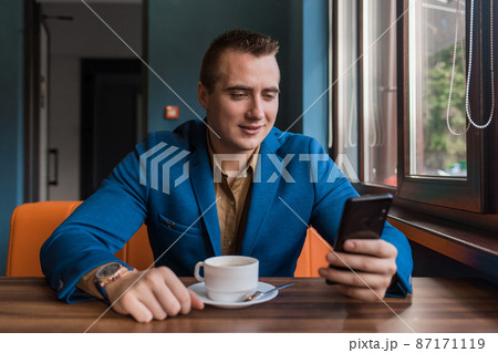 A young satisfied handsome guy businessman of European appearance portrait, uses a smartphone or mobile phone sitting in a cafe at a table on a coffee break 87171119