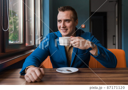 A stylish, positive businessman of Caucasian appearance, a man in a jacket and shirt sits at a table in a cafe on a coffee break and looks away or out the window 87171120