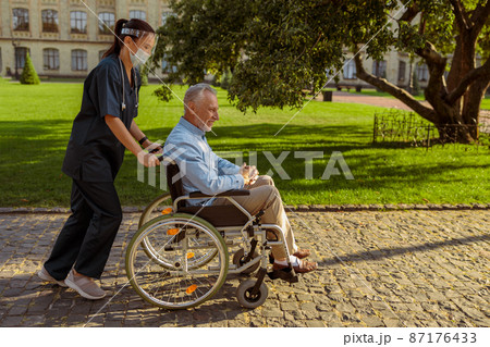 Full length shot of caring nurse with senior man in wheelchair on a walk in the park near hospital Full length shot of caring nurse with senior man in wheelchair on a walk in the park near hospital 87176433