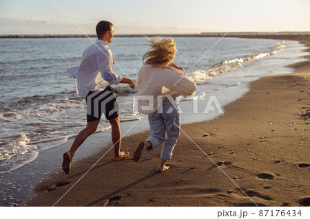 Back photo of man and woman running holding hands along the ocean Back photo of man and woman running holding hands along the ocean 87176434