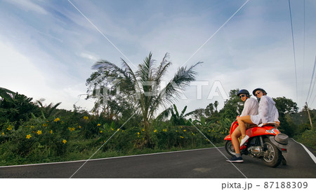 Scooter road trip. Love couple on red motorbike in white clothes on forest road trail. Just married woman and man kiss, hugs, sit on motorbike. 87188309