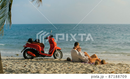 Scooter road trip. Lovely couple on red motorbike in white clothes on sand beach. People walking near the tropical palm trees, sea. Motorcycle rent. 87188709