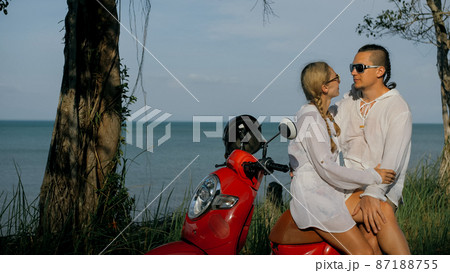 Love couple on red motorbike in white clothes on forest road trail trip. Two caucasian tourist woman and man kiss hugs sit on scooter. Love couple on red motorbike in white clothes on forest road trail trip. Two caucasian tourist woman and man kiss hugs sit on scooter. 87188755