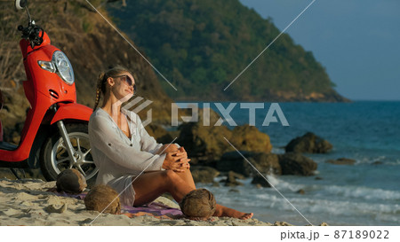 Scooter road trip. Woman alone on red motorbike in white clothes on sand beach by ocean. One girl caucasian tourist walk near tropical palm tree. 87189022