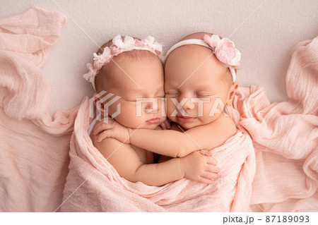 Tiny newborn twin girls. A newborn twin sleeps next to his sister. Newborn twin girls on the background of a pink blanket with pink bandages. The girls gently hug and kiss their sister in a cute pose. 87189093