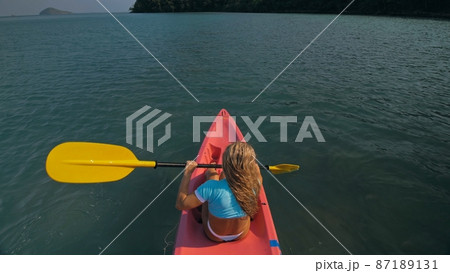 Seductive blonde woman in blue swimsuit sails on pink plastic kayak along azure sea at exotic resort backside upper above view. 87189131