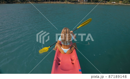 Seductive blonde woman in blue swimsuit sails on pink plastic kayak along azure sea at exotic resort backside upper above view. 87189148