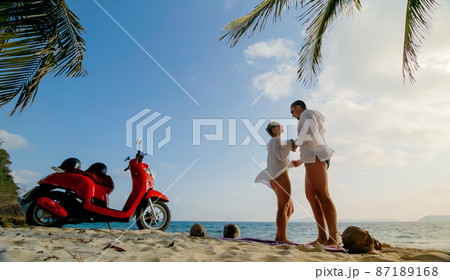 Scooter road trip. Lovely couple on red motorbike in white clothes on sand beach. People walking near the tropical palm trees, sea. Motorcycle rent. Scooter road trip. Lovely couple on red motorbike in white clothes on sand beach. People walking near the tropical palm trees, sea. Motorcycle rent. 87189168