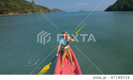 Pretty sportive blonde lady in stylish swimsuit sails on pink plastic canoe along azure ocean bay at exotic resort. Pretty sportive blonde lady in stylish swimsuit sails on pink plastic canoe along azure ocean bay at exotic resort. 87189258
