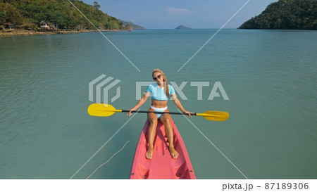 Pretty sportive blonde lady in stylish swimsuit sails on pink plastic canoe along azure ocean bay at exotic resort. 87189306