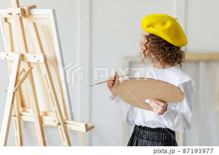 Portrait of little girl artist in yellow beret who paints picture in studio while holds pallete Portrait of little girl artist in yellow beret who paints picture in studio while holds pallete 87193977