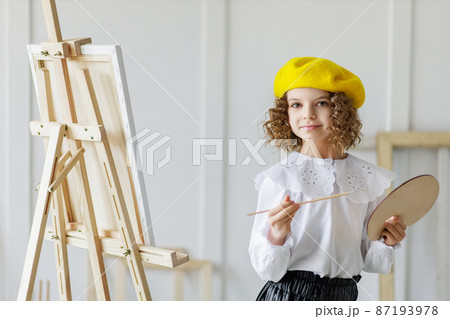 Portrait of little girl artist in yellow beret who paints picture in studio while holds pallete Portrait of little girl artist in yellow beret who paints picture in studio while holds pallete 87193978