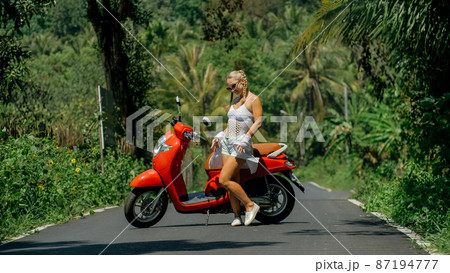 Woman on red scooter in white clothes drive on forest road trail. Dancing trip. One girl caucasian tourist in sunglasses dance, relax near motorbike. Woman on red scooter in white clothes drive on forest road trail. Dancing trip. One girl caucasian tourist in sunglasses dance, relax near motorbike. 87194777