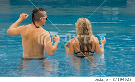 The loving couple hugs and kisses, drinking blue cocktail alcohol liquor in swimming pool at hotel outdoor. Portrait of caucasian man and woman. The loving couple hugs and kisses, drinking blue cocktail alcohol liquor in swimming pool at hotel outdoor. Portrait of caucasian man and woman. 87194928