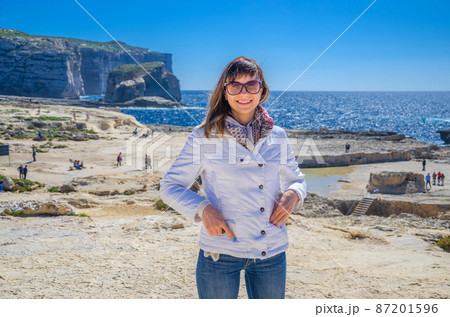 Young woman traveler with white jacket and sunglasses looking at camera and smile on coast of Dwejra Bay 87201596