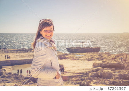 Young woman traveler with white jacket and sunglasses looking at camera and smile on coast of Dwejra Bay Young woman traveler with white jacket and sunglasses looking at camera and smile on coast of Dwejra Bay 87201597