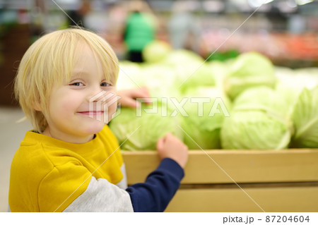 Little boy boy in a food store or a supermarket choosing fresh organic cabbage. Healthy food for family with kids Little boy boy in a food store or a supermarket choosing fresh organic cabbage. Healthy food for family with kids 87204604