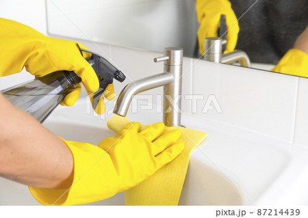 Close-up of male hands in yellow rubber gloves holding sponge and spray with cleaning agent, maid washing and polishing sink in bathroom 87214439