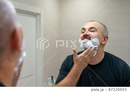 Portrait of handsome gray-haired middle-aged man applying shaving foam to trim his beard Portrait of handsome gray-haired middle-aged man applying shaving foam to trim his beard 87220055