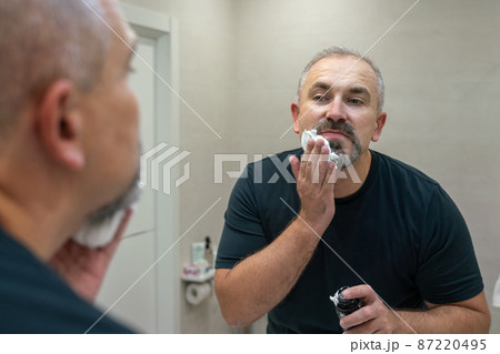 Portrait of handsome gray-haired middle-aged man applying shaving foam to trim his beard Portrait of handsome gray-haired middle-aged man applying shaving foam to trim his beard 87220495