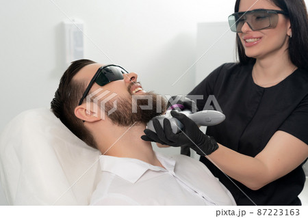 Young bearded man getting laser facial treatment by professional cosmetologist in a beauty clinic. Healthy man lifestyle concept Young bearded man getting laser facial treatment by professional cosmetologist in a beauty clinic. Healthy man lifestyle concept 87223163