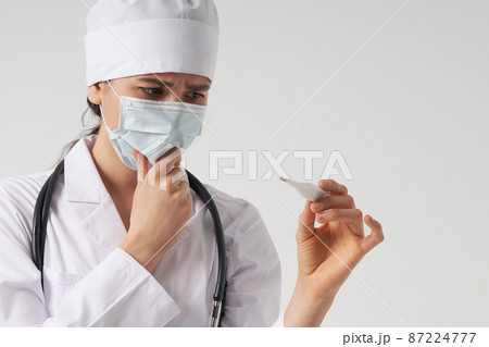 Doctor wearing face mask looking at measurement on medical thermometer over white background with copy space 87224777