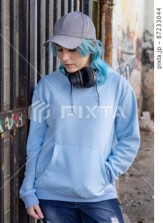 Blue haired teenage girl in light blue hoodie with headphones stays near a gate 87233044