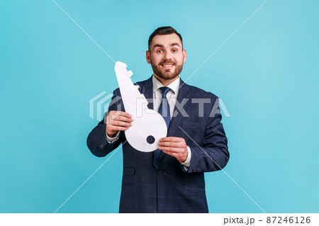 Portrait of man wearing official style suit holding in hand big paper key as symbol of her own apartment, dreams comes true, state housing program. Indoor studio shot isolated on blue background. Portrait of man wearing official style suit holding in hand big paper key as symbol of her own apartment, dreams comes true, state housing program. Indoor studio shot isolated on blue background. 87246126