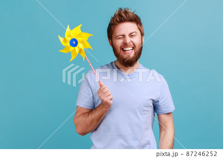 Portrait of positive delighted handsome bearded man smiling carefree and winking to camera, holding paper windmill, pinwheel toy on stick. Indoor studio shot isolated on blue background. Portrait of positive delighted handsome bearded man smiling carefree and winking to camera, holding paper windmill, pinwheel toy on stick. Indoor studio shot isolated on blue background. 87246252