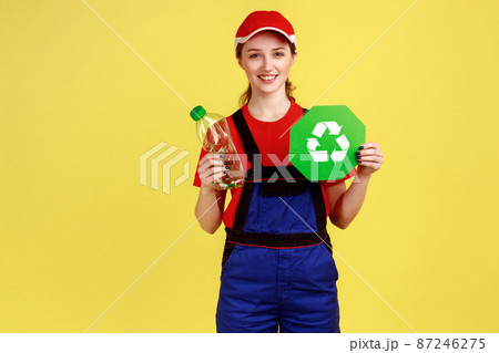 Smiling positive worker woman volunteer standing with empty plastic bottle and green recycling sign, wearing overalls and red cap. Indoor studio shot isolated on yellow background. Smiling positive worker woman volunteer standing with empty plastic bottle and green recycling sign, wearing overalls and red cap. Indoor studio shot isolated on yellow background. 87246275