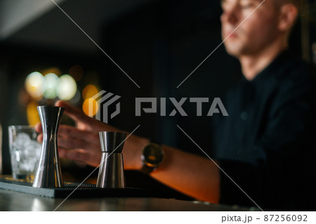 Low-angle view of professional bartender preparing refreshing alcoholic cocktail standing behind bar counter, focus in foreground to metal jigger, in modern nightclub with dark interior. Low-angle view of professional bartender preparing refreshing alcoholic cocktail standing behind bar counter, focus in foreground to metal jigger, in modern nightclub with dark interior. 87256092