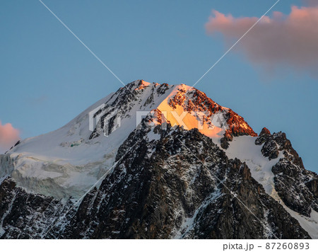 Top black rocks in evening golden sunshine and white-snow pointy peak. Minimalistic atmospheric landscape with high snowy mountain with peaked top under cirrus clouds in sky. Top black rocks in evening golden sunshine and white-snow pointy peak. Minimalistic atmospheric landscape with high snowy mountain with peaked top under cirrus clouds in sky. 87260893
