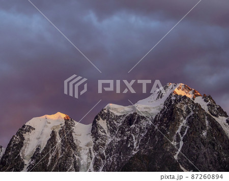 Red fiery cloud in the evening over the silhouette of sharp mountains. Top black rocks in evening golden sunshine and white-snow pointy peak. Magnificent purple sunset in the mountains. Red fiery cloud in the evening over the silhouette of sharp mountains. Top black rocks in evening golden sunshine and white-snow pointy peak. Magnificent purple sunset in the mountains. 87260894