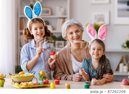 Loving grandmother teaching happy little kids girls to decorate Easter eggs while sitting in kitchen Loving grandmother teaching happy little kids girls to decorate Easter eggs while sitting in kitchen 87262206