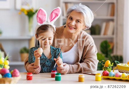 Loving grandmother teaching happy little kid girl to decorate Easter eggs while sitting in kitchen Loving grandmother teaching happy little kid girl to decorate Easter eggs while sitting in kitchen 87262209