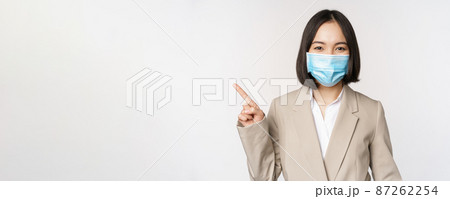 Coronavirus and work concept. Portrait of woman in medical face mask, pointing finger left, showing logo or banner, advertisement, white background Coronavirus and work concept. Portrait of woman in medical face mask, pointing finger left, showing logo or banner, advertisement, white background 87262254