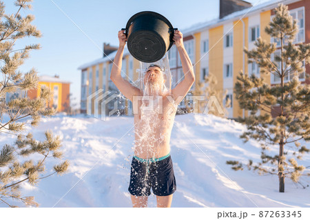 Front view of young strong-willed man tempering body, pouring water onto himself, developing resistance to cold outdoors, enjoying winter freshness outdoors in frozen sunny day. 87263345